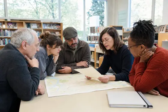 Five adults around a library table with a large city map, one reading case notes aloud