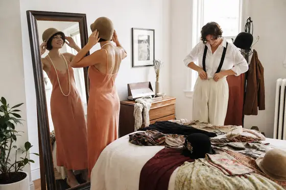 Two friends try on a cloche hat, pearls and suspenders in front of a mirror
